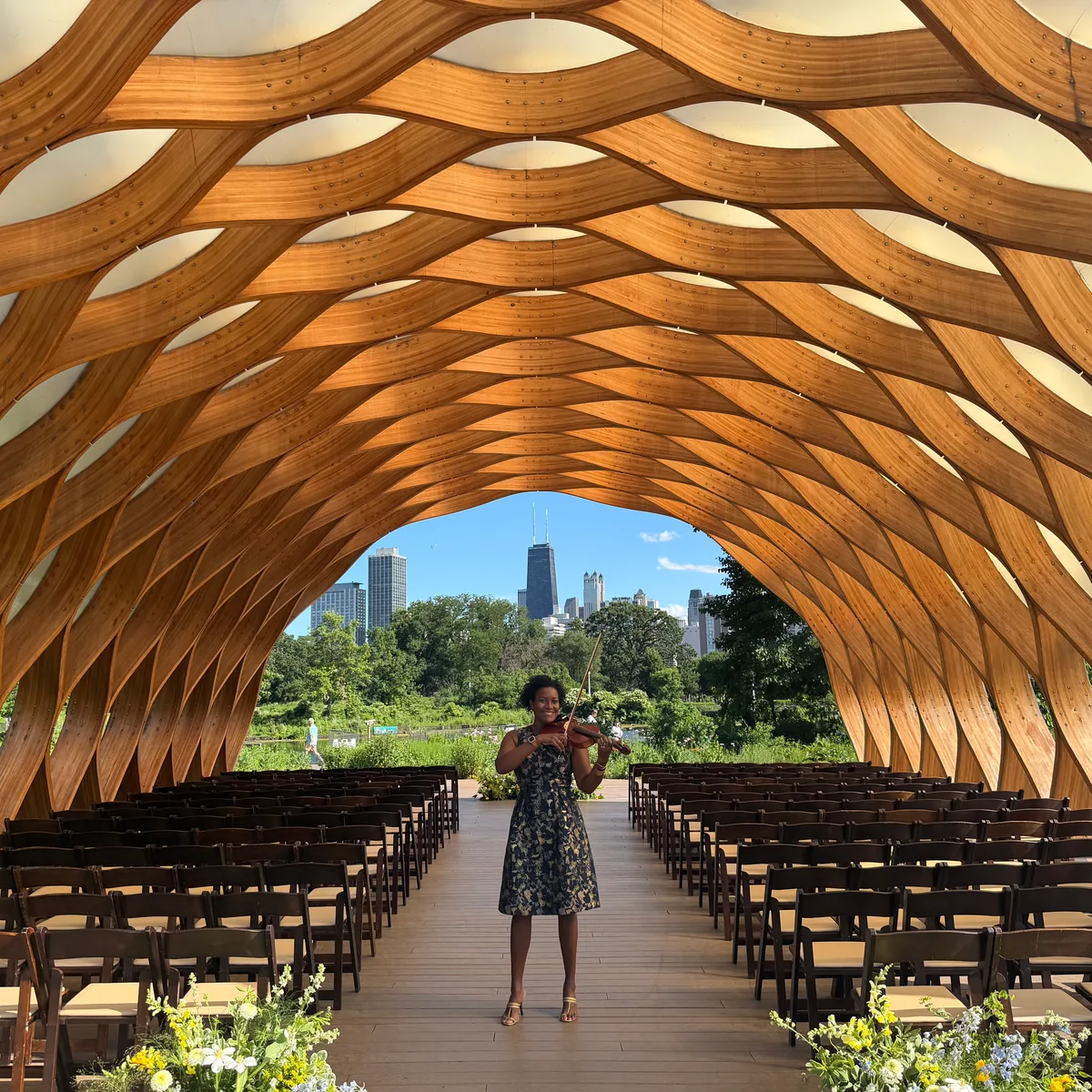 Adia performing violin beneath a wooden honeycomb pavilion, with ceremony seating and the Chicago skyline behind her