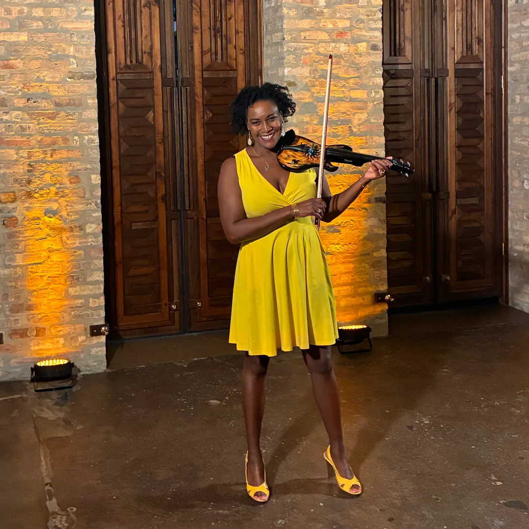 Adia smiling with her electric violin in a yellow dress before tall wooden doors