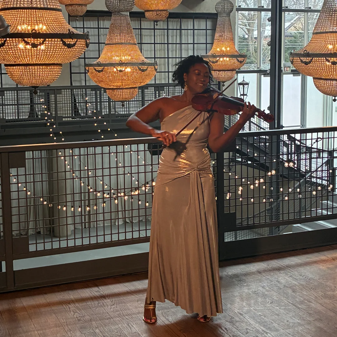 Adia performing violin in a gold gown beneath crystal chandeliers